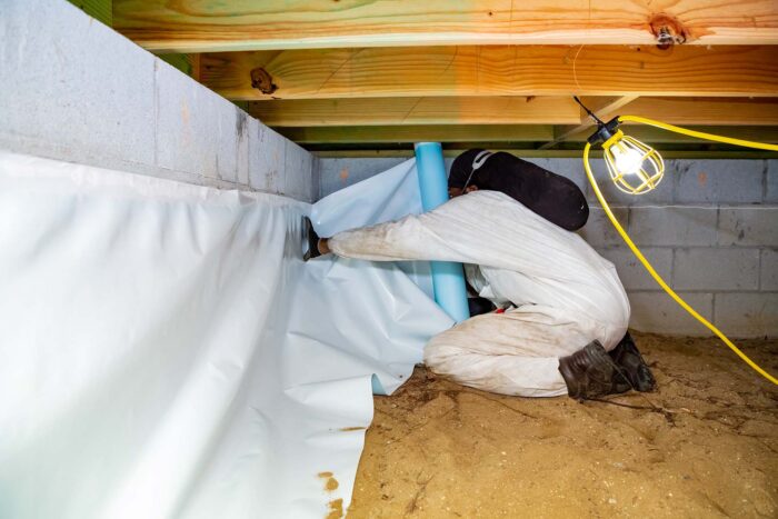 A person in protective clothing installs a plastic vapor barrier for crawl space mold removal along the wall, illuminated by a hanging work light.