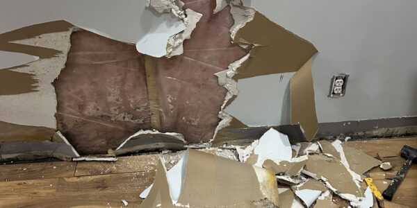 A wall with a large hole exposing insulation and studs, surrounded by broken drywall pieces scattered on a wooden floor. Tools are visible near the debris, hinting at repairs to check for hidden mold behind baseboards near the electrical outlet.