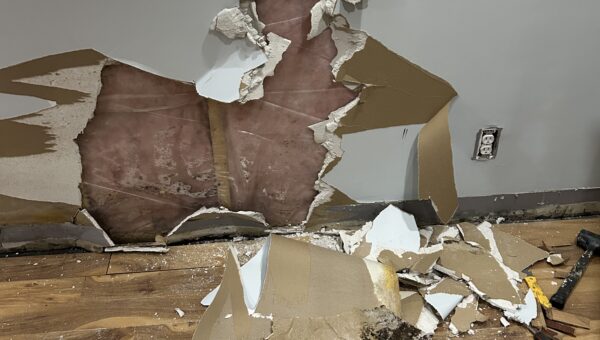 A wall with a large hole exposing insulation and studs, surrounded by broken drywall pieces scattered on a wooden floor. Tools are visible near the debris, hinting at repairs to check for hidden mold behind baseboards near the electrical outlet.