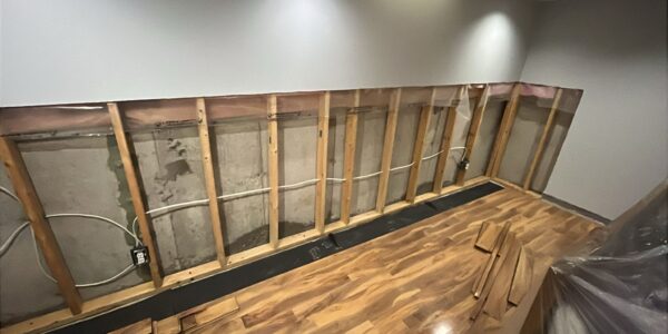 A room with wall studs exposed, drywall partially removed, and electrical wiring visible. The floor has wood panels, some stacked on the side. Ceiling lights illuminate the area—possible hidden mold behind baseboards is revealed during renovation.