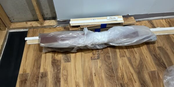 A bundle of wooden planks wrapped in plastic lies on a wood-patterned floor, next to stacked trim pieces and an unfinished wall with exposed framing—ideal for checking for hidden mold behind baseboards during installation.