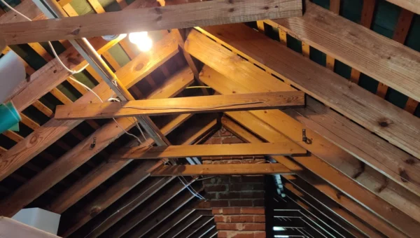 Wooden attic with exposed beams, a brick chimney running through the center, plastic storage bins, and miscellaneous household items scattered around. A single lightbulb is on, highlighting the need for proper attic ventilation Toronto homes require.