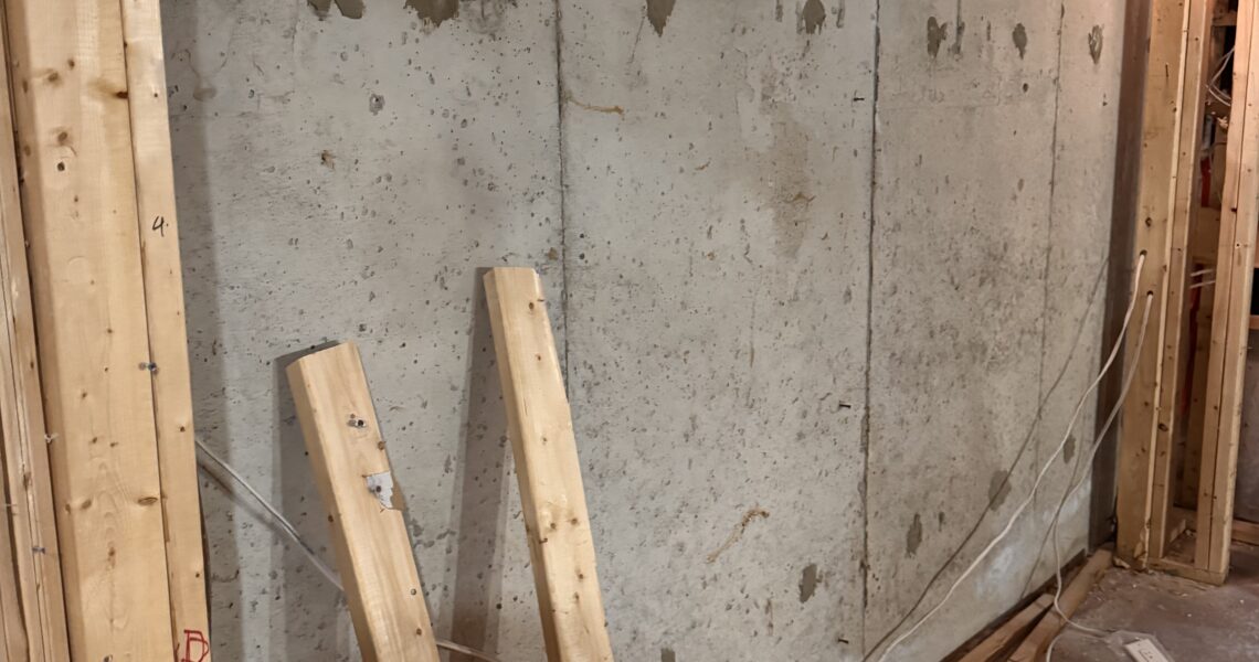 Unfinished basement with exposed wooden framing, bare concrete wall, loose electrical wires, and two wooden beams leaning against the wall. Some scattered construction materials are on the ground.