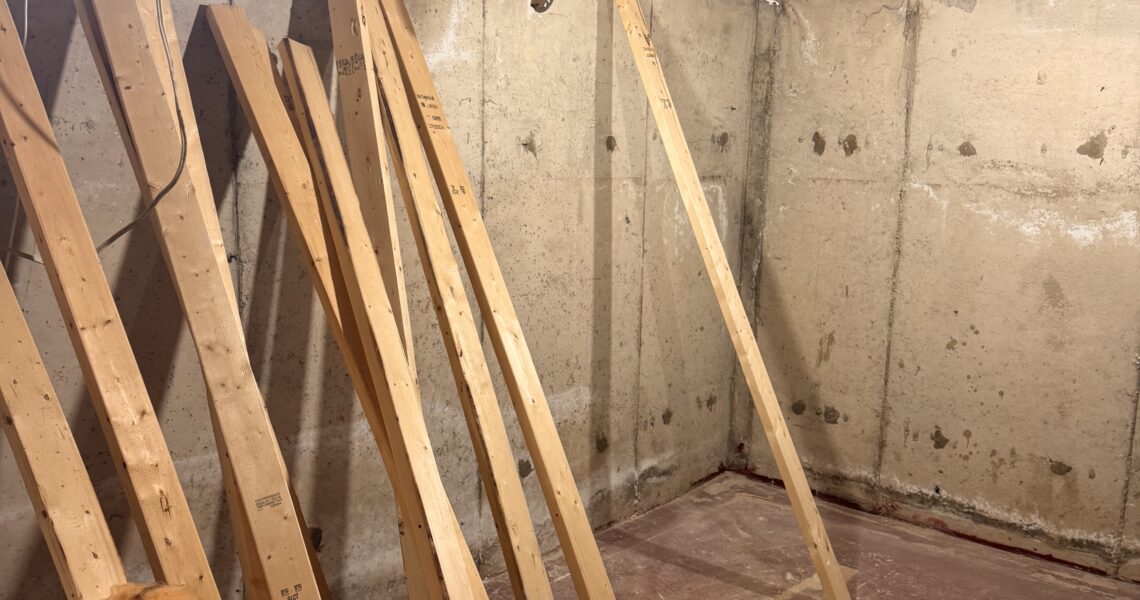 Unfinished basement with exposed wooden ceiling beams, concrete walls, upright wooden studs leaning against the wall, exposed insulation, loose wires, and a single hanging light bulb.