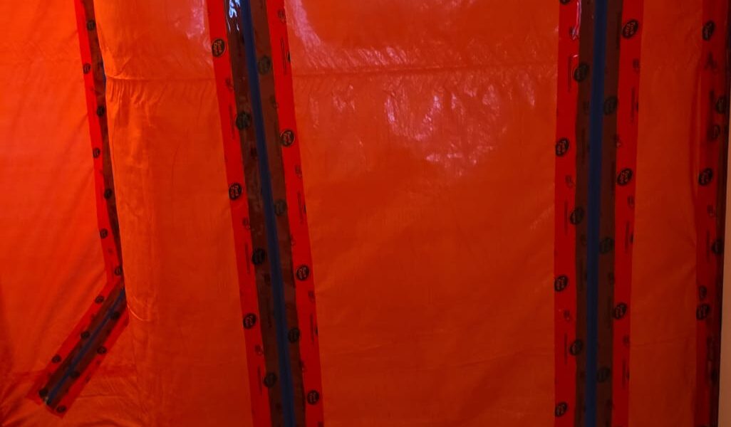 A doorway and wall covered with bright orange plastic sheeting, sealed with red and blue tape, likely creating a barrier for mold remediation in Richmond Hill or restoration work. A brown tiled floor is visible at the bottom.
