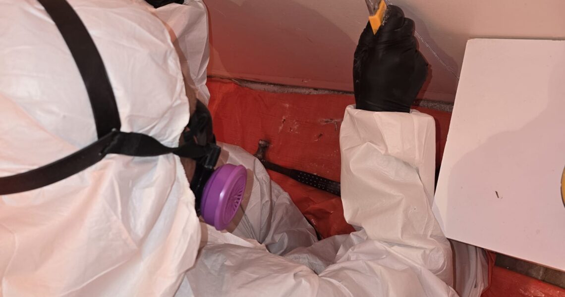 A person in protective gear and gloves uses a utility knife to cut into a white ceiling during mold remediation in Richmond Hill. The floor is covered with a red plastic sheet, and a purple respirator mask is worn for safety.