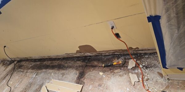 A room under renovation with the baseboards removed, exposing damaged drywall and old flooring—possible signs for mold remediation Richmond Hill. Debris, tools, and an orange extension cord are scattered on the floor; part of the wall is covered in plastic and blue tape.