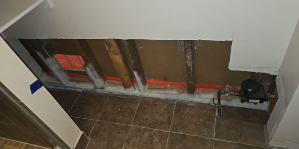 A section of drywall has been removed from the bottom of a wall, exposing wooden studs, pipes, insulation, and a water meter—common sights during mold remediation in Richmond Hill. The surrounding floor is tiled.