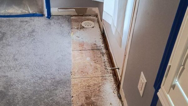 A section of carpet near the balcony door has been removed, exposing stained plywood subfloor—possibly from mold. Nearby walls and baseboards are taped off with blue painters tape, suggesting ongoing renovation or repair work in this Oakville home.