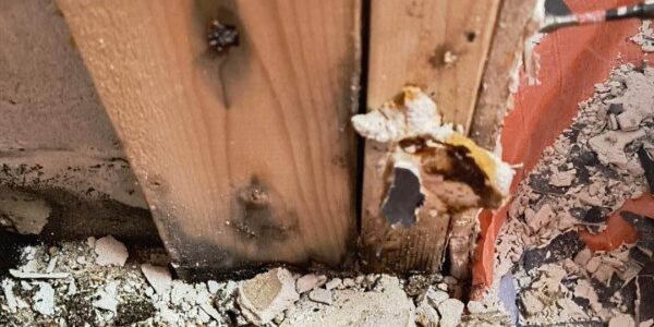 A close-up of a damaged wooden wall stud with a hole and debris on the floor, including broken plaster and pieces of wood, next to an orange and blue tarp.
