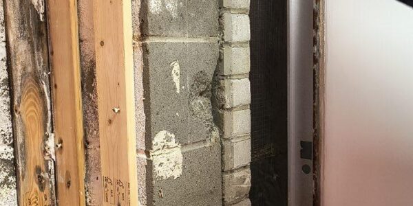 A person wearing a blue glove and hat exposes an interior wall section, revealing bricks, cinder blocks, and wooden framing next to construction equipment.