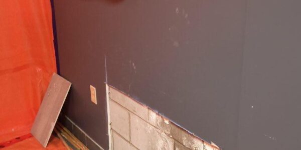 A section of drywall has been removed from a gray wall, exposing concrete blocks underneath. Debris and broken drywall pieces are scattered on an orange tarp on the floor. A covered item is mounted on the wall above.