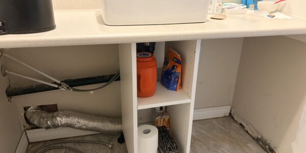 Under-sink cabinet with cleaning supplies, paper towels, and exposed pipes. Countertop holds a sink, soap, and toiletries; a child is visible in the mirror taking a photo. The area appears to be under renovation for Mold Remediation Oakville.