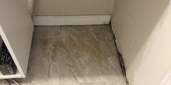 A laundry area with two electrical outlets, an exposed dryer vent hose, and gray tile flooring. Dark mold or water damage is visible along the bottom edge of the wall and baseboard, indicating a need for Mold Remediation Oakville services.