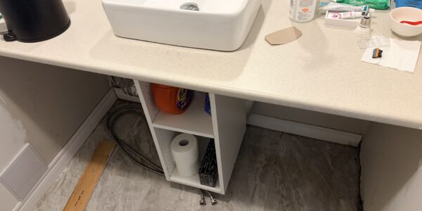 A bathroom sink with a countertop, soap, disinfectant, wipes, and a black trash can. Under the sink are exposed pipes, detergent, paper towels, and a loose wooden plank—an ideal spot for Mold Remediation Oakville to address potential concerns.