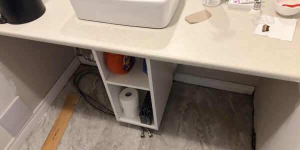 A bathroom sink with a cluttered countertop, including bottles, soap, and wipes. Below the sink is an open shelf with toilet paper and cleaning products—ideal for Mold Remediation Oakville if exposed pipes and moisture become an issue.