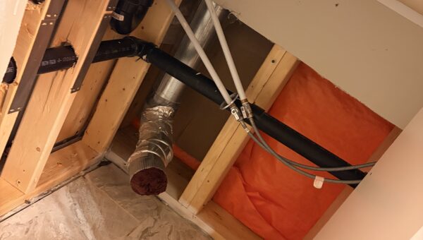 Exposed plumbing and pipes under a kitchen sink, with cabinet panels removed, revealing wood framing, insulation, ductwork, and floor tiles—ideal conditions where Mold Remediation Oakville may be necessary.