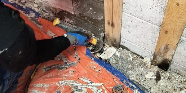 A person wearing a blue cap and gloves removes damaged drywall near a wall, exposing wooden studs. Debris, tools, and a power drill are scattered on an orange tarp on the floor.