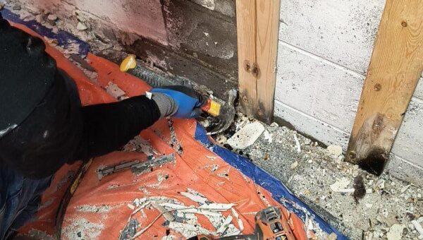 A person wearing a blue cap and gloves removes damaged drywall near a wall, exposing wooden studs. Debris, tools, and a power drill are scattered on an orange tarp on the floor.