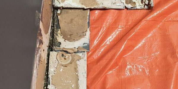 Several damaged and peeling rectangular tiles are set against a dark wall, with an orange tarp covering the floor nearby, suggesting ongoing renovation or repair work.