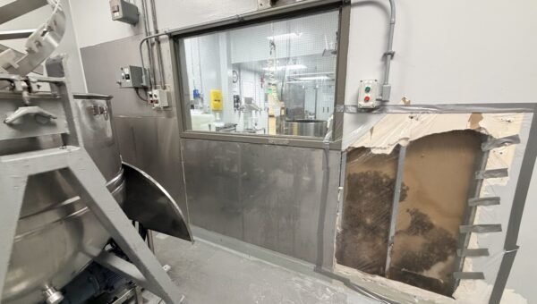 A room with industrial equipment, a large window revealing another room, and a damaged wall section exposing moldy insulation—an environment that highlights the need for professional commercial mould remediation in Cambridge, Ontario.
