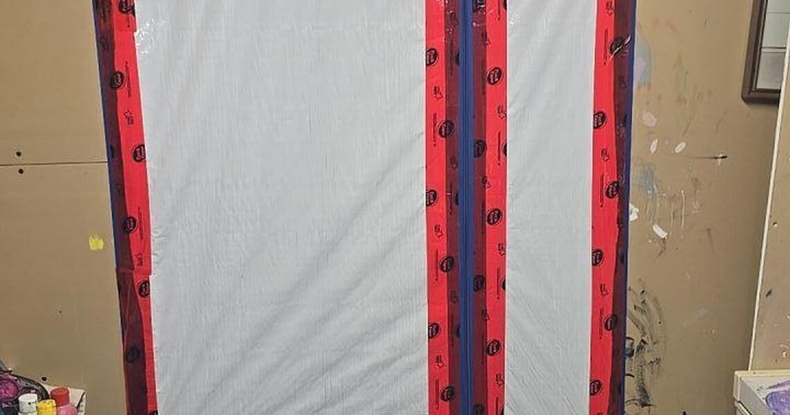 A plastic sheet taped over a doorway with red and black tape forming a large, angular J shape. The setting appears to be a cluttered room or basement with paint stains and various supplies nearby.